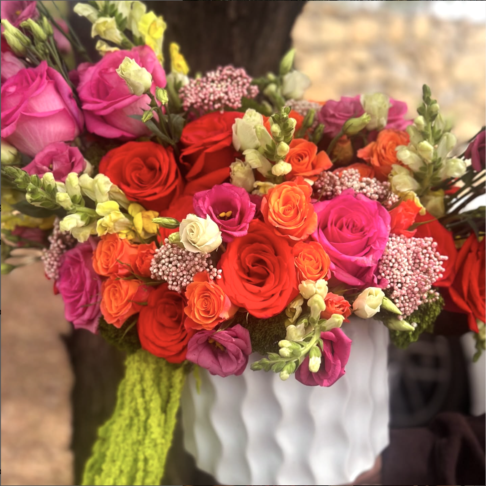 Ramo de flores variadas en un jarrón blanco con textura, con rosas rojas, rosas rosadas, claveles blancos, y otros pequeños flores en tonos amarillos, rosados y naranjas.