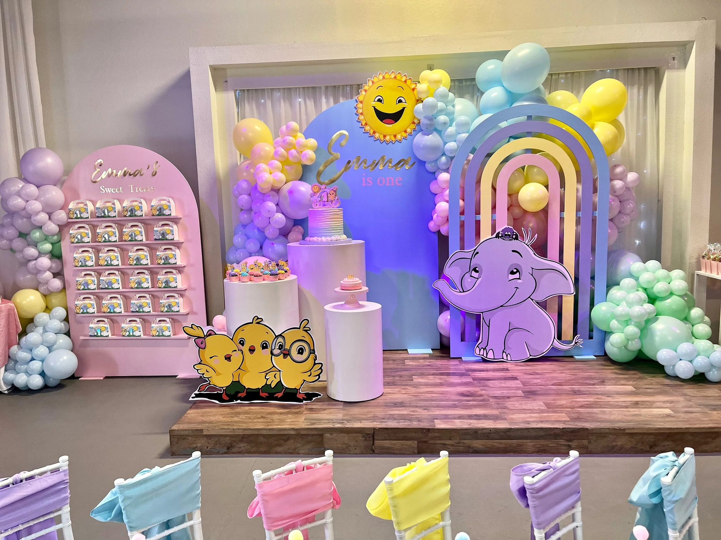Decorated birthday party setup with colorful balloons, a pink display with candy, cartoon character cutouts, a cake, and a backdrop featuring a rainbow, a smiling sun, and an elephant, celebrating Emma's first birthday.