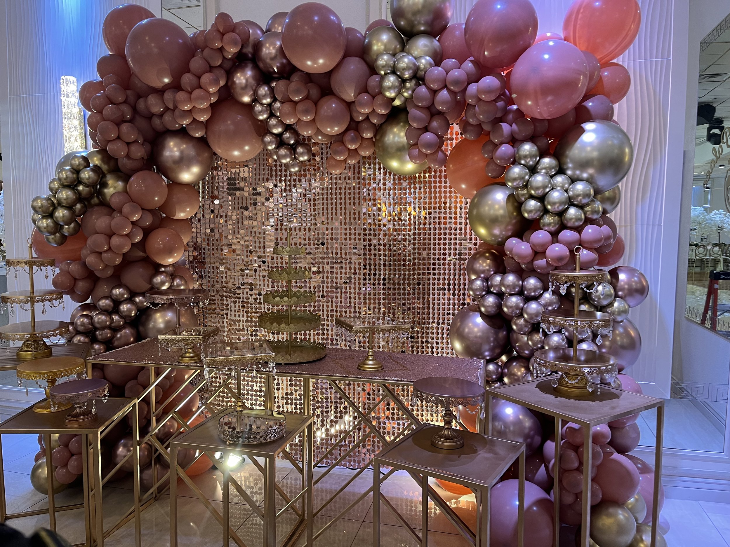 Decorative balloon arch in shades of pink, gold, and rose with a glittery backdrop and gold tables with tiered trays.