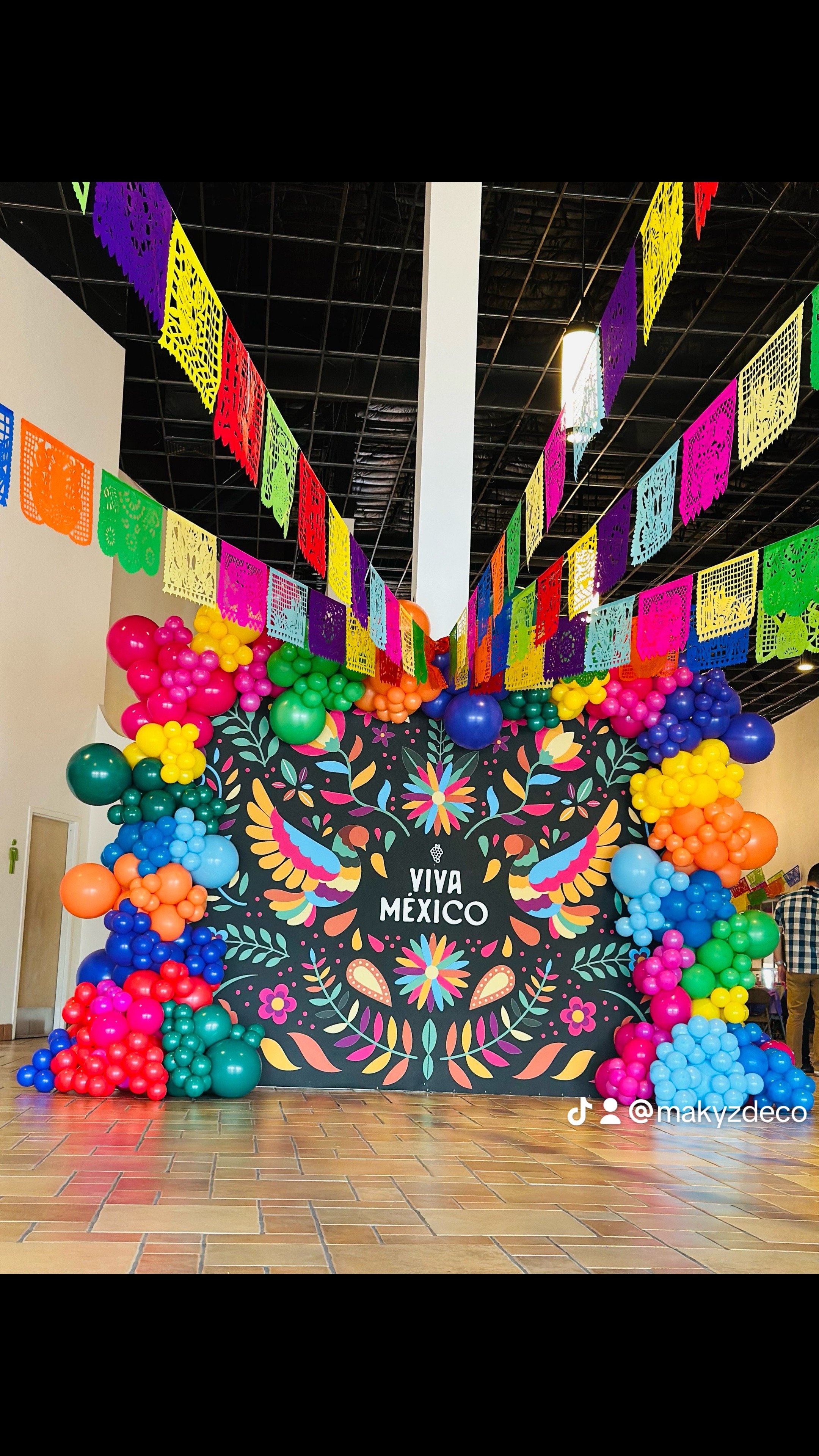 Colorful decorations for a Mexican celebration including balloons and papel picado banners with the text "Viva México" on a floral mural background.