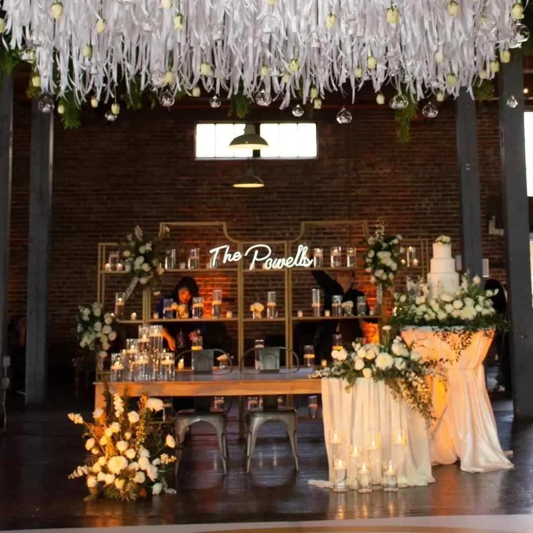 Elegant wedding reception setup with floral arrangements, candles, and a wedding cake. A neon sign reads 'The Powells' behind a table decorated with flowers and candles.
