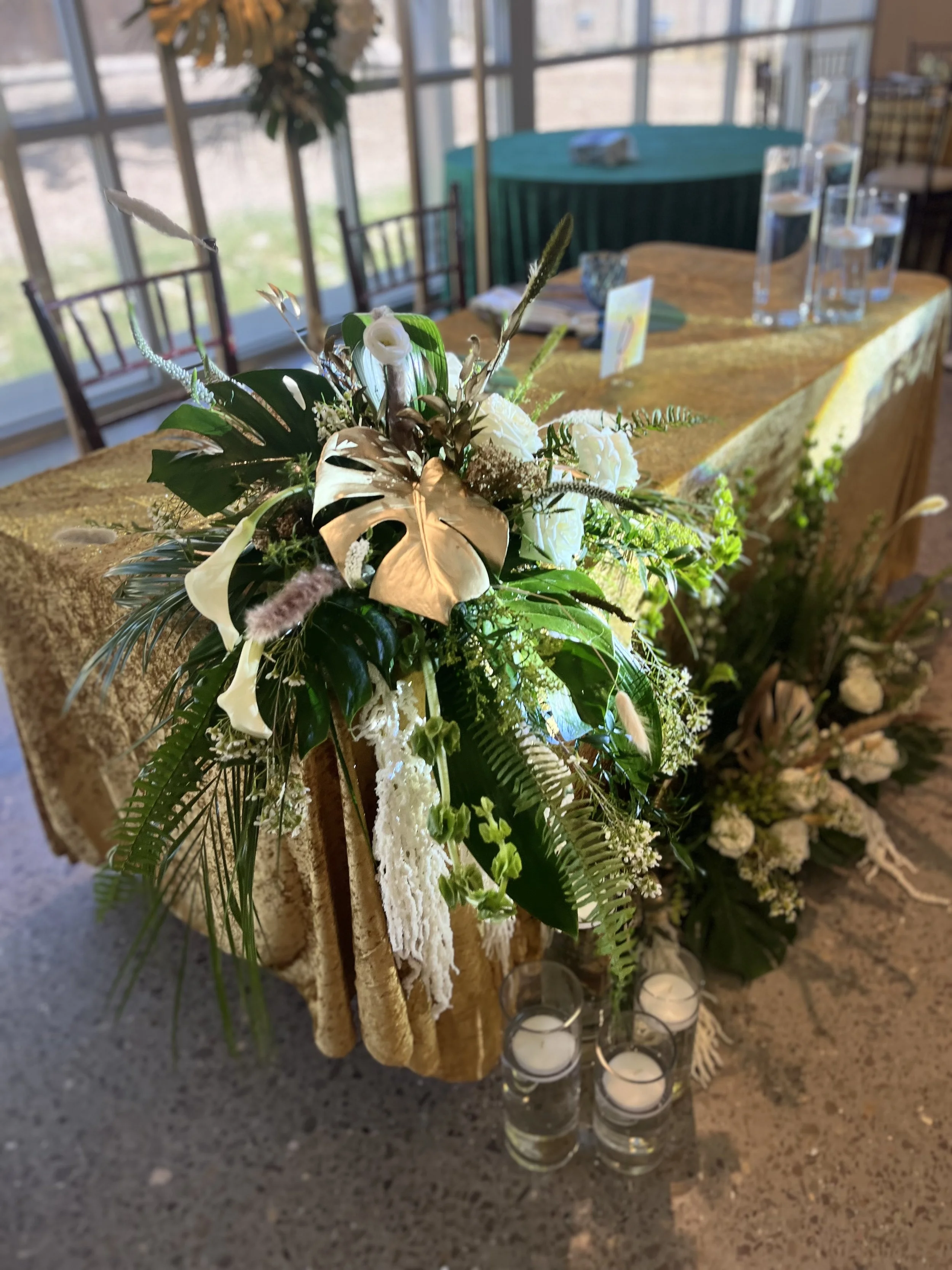 A table decorated with a gold tablecloth, featuring a green and white floral centerpiece, surrounded by candles in glass holders, set in a bright room with large windows.