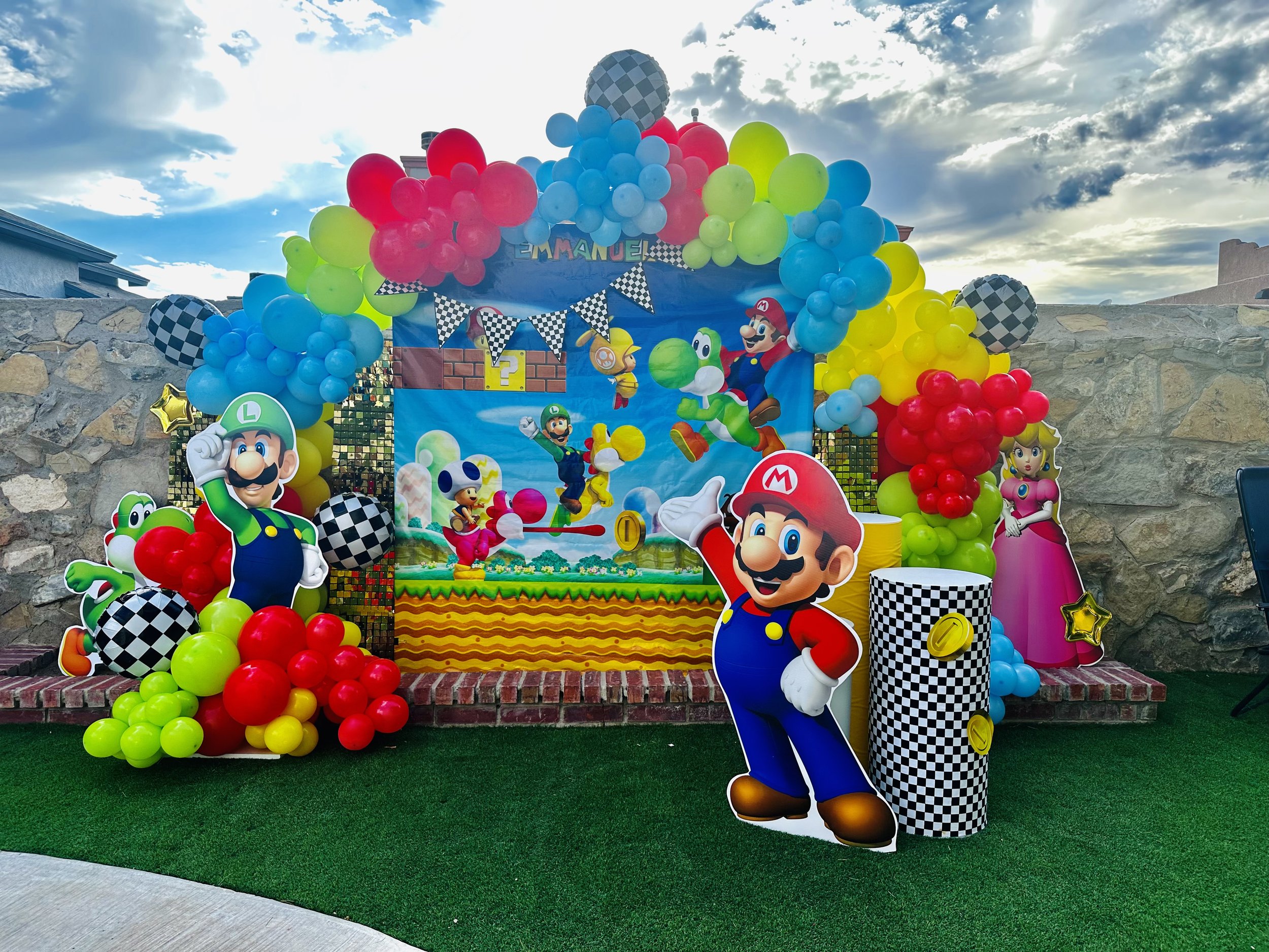 Super Mario themed birthday party setup with colorful balloons, cardboard cutouts of Mario, Luigi, Princess Peach, Toad, and Yoshi, and a backdrop depicting a Super Mario game scene.