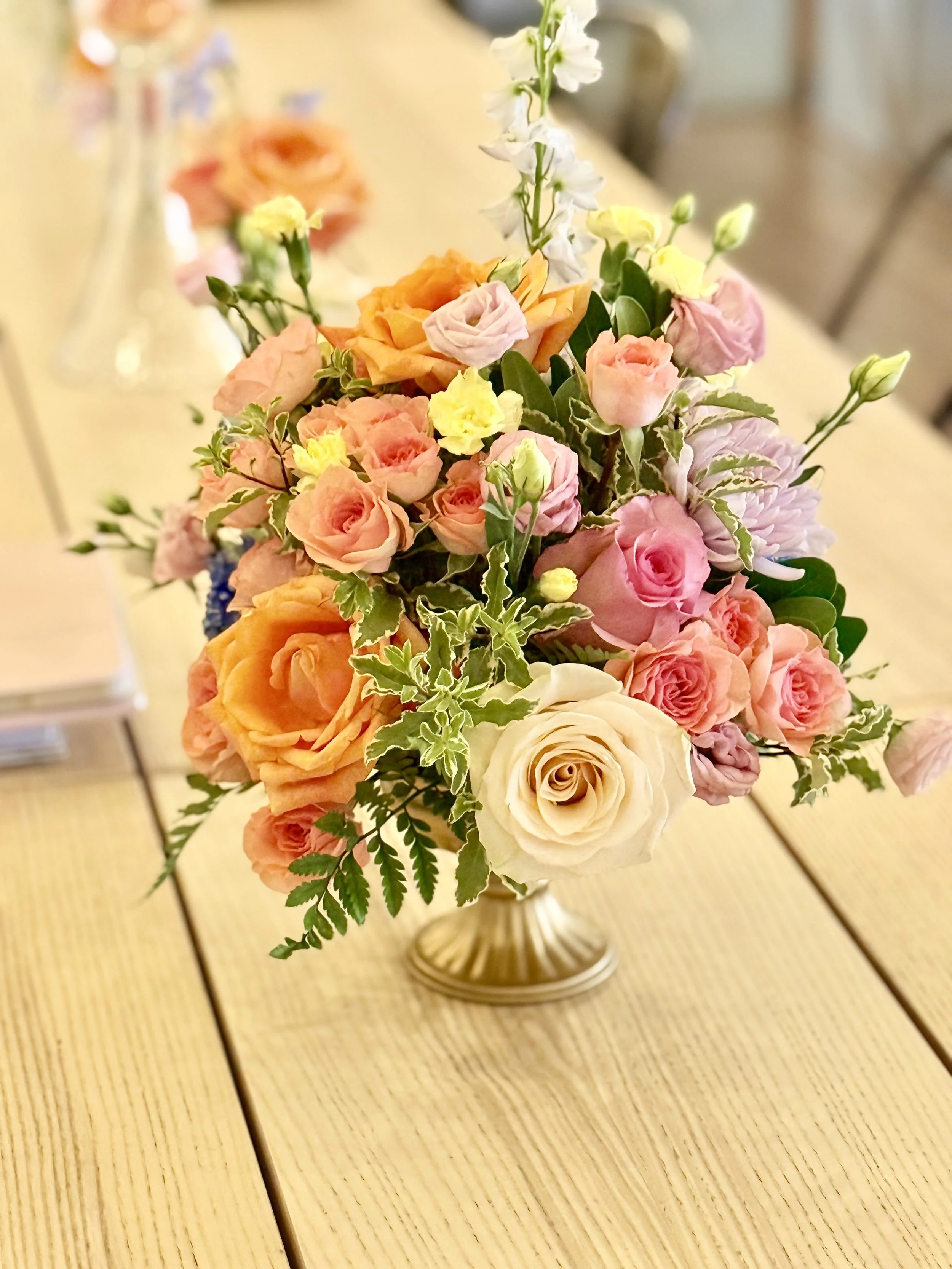 Colorful bouquet of roses, carnations, and greenery in a gold vase on a wooden table.