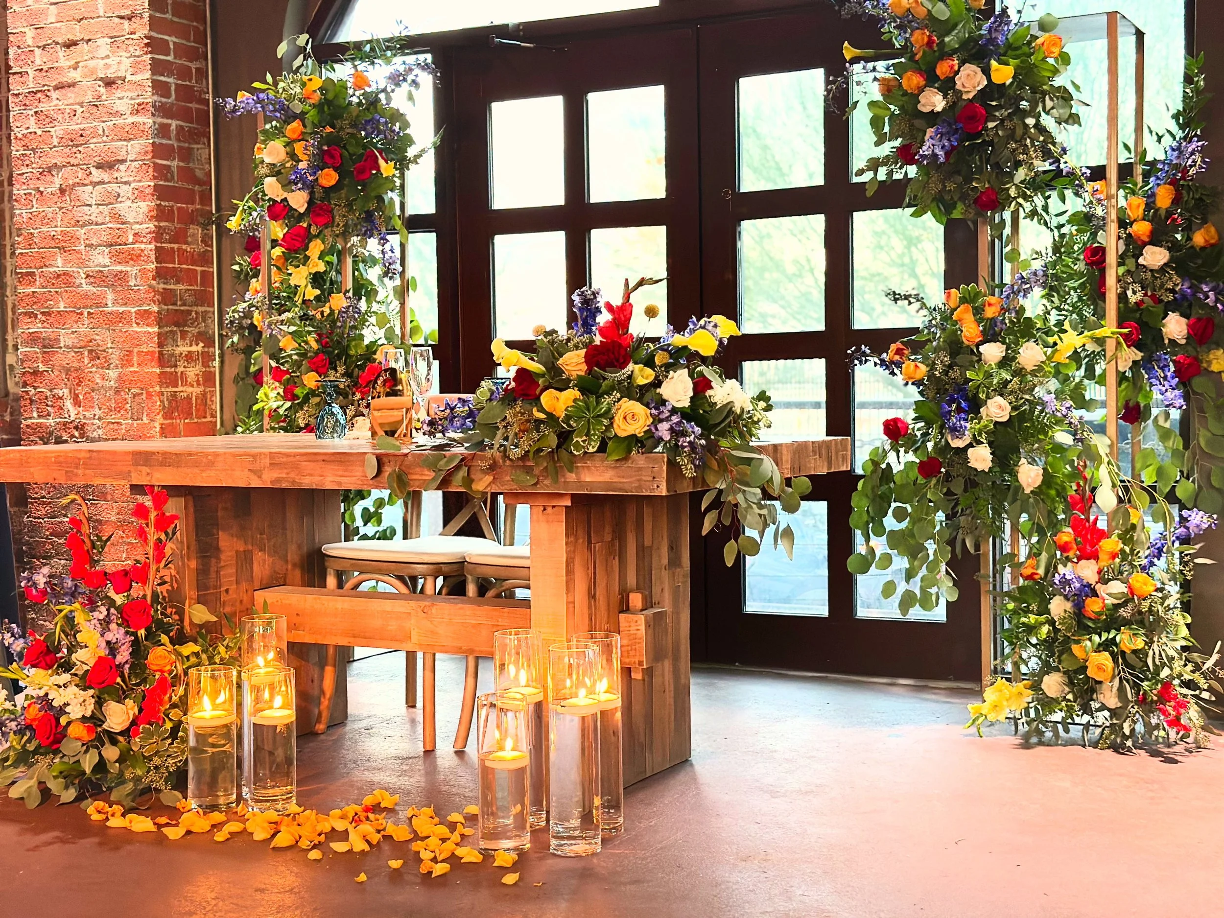 A floral wedding altar decorated with colorful flowers, flowers on the floor, and candles in glass holders, set inside a room with brick and glass walls.