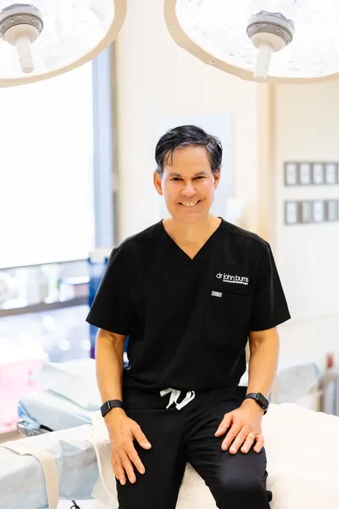 Dr. John L. Burns, Jr., MD in black scrubs sitting on an examination table in a medical clinic or hospital, with medical equipment and window in the background.