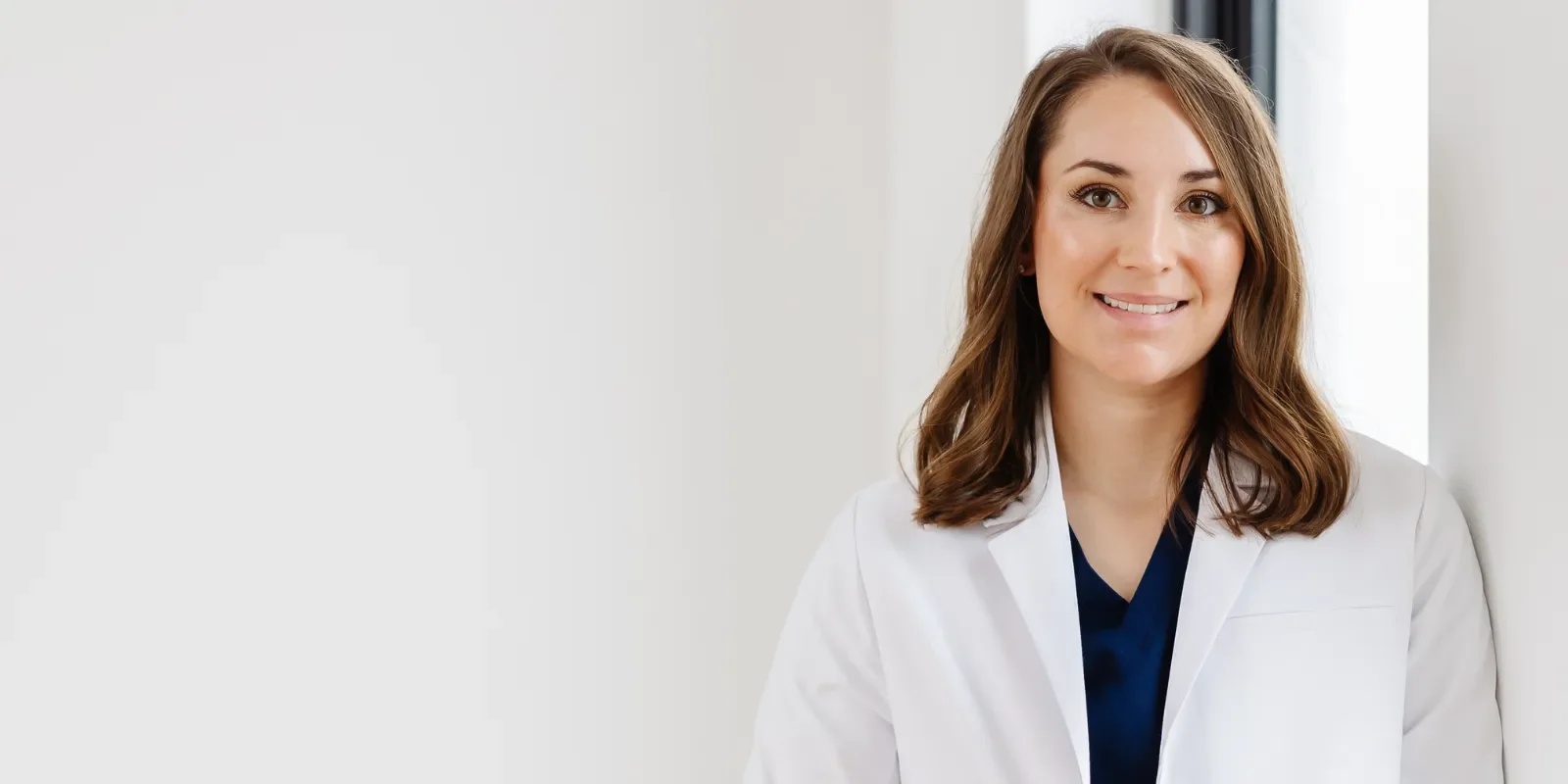 Nicole Stennet, PA-C with shoulder-length brown hair, wearing a white lab coat, looking at the camera near a window in a bright, modern medical or laboratory setting.
