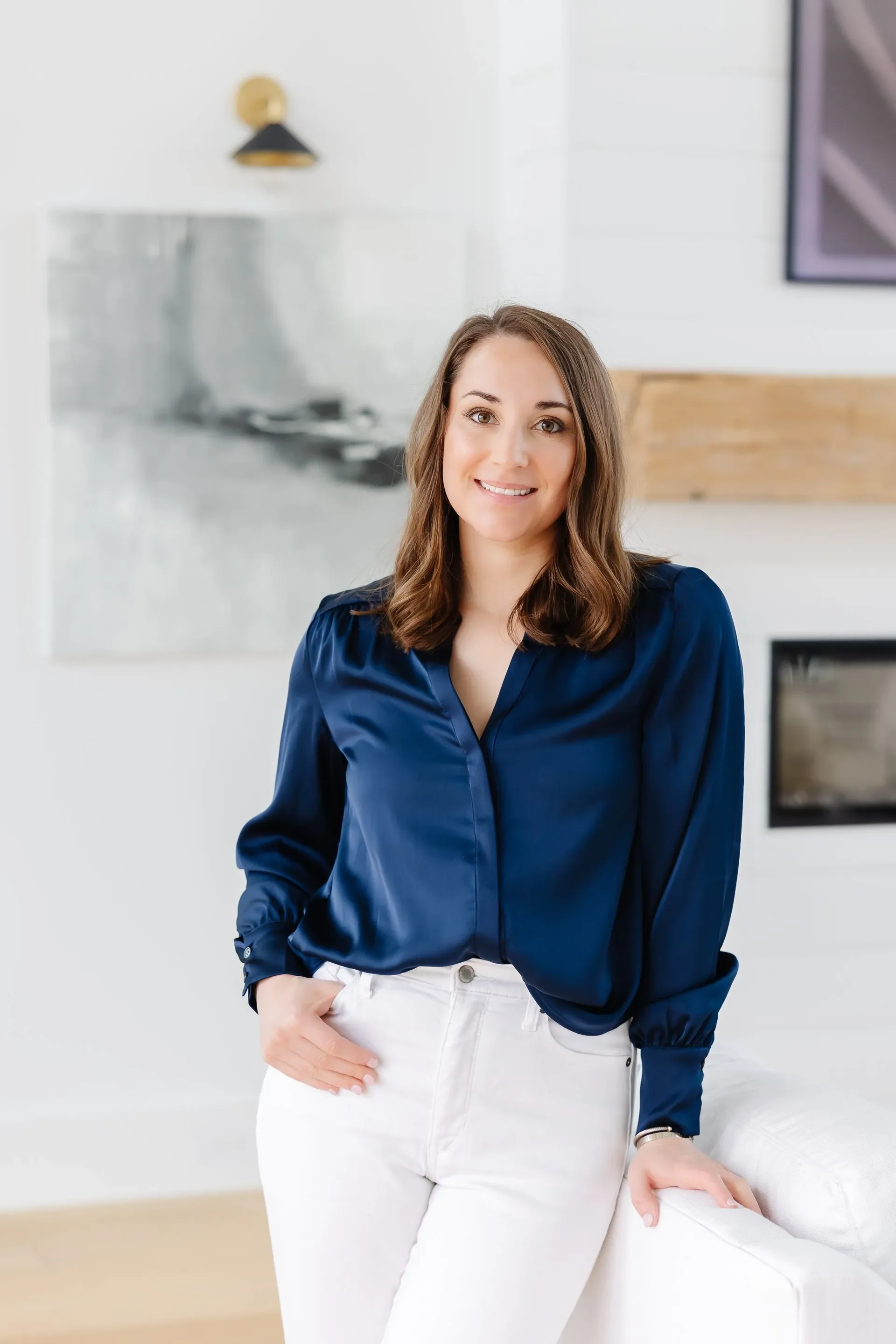 Nicole Stennet, PA-C with shoulder-length brown hair wearing a navy blue satin blouse and white pants, smiling in a modern living room with abstract artwork and a fireplace in the background.