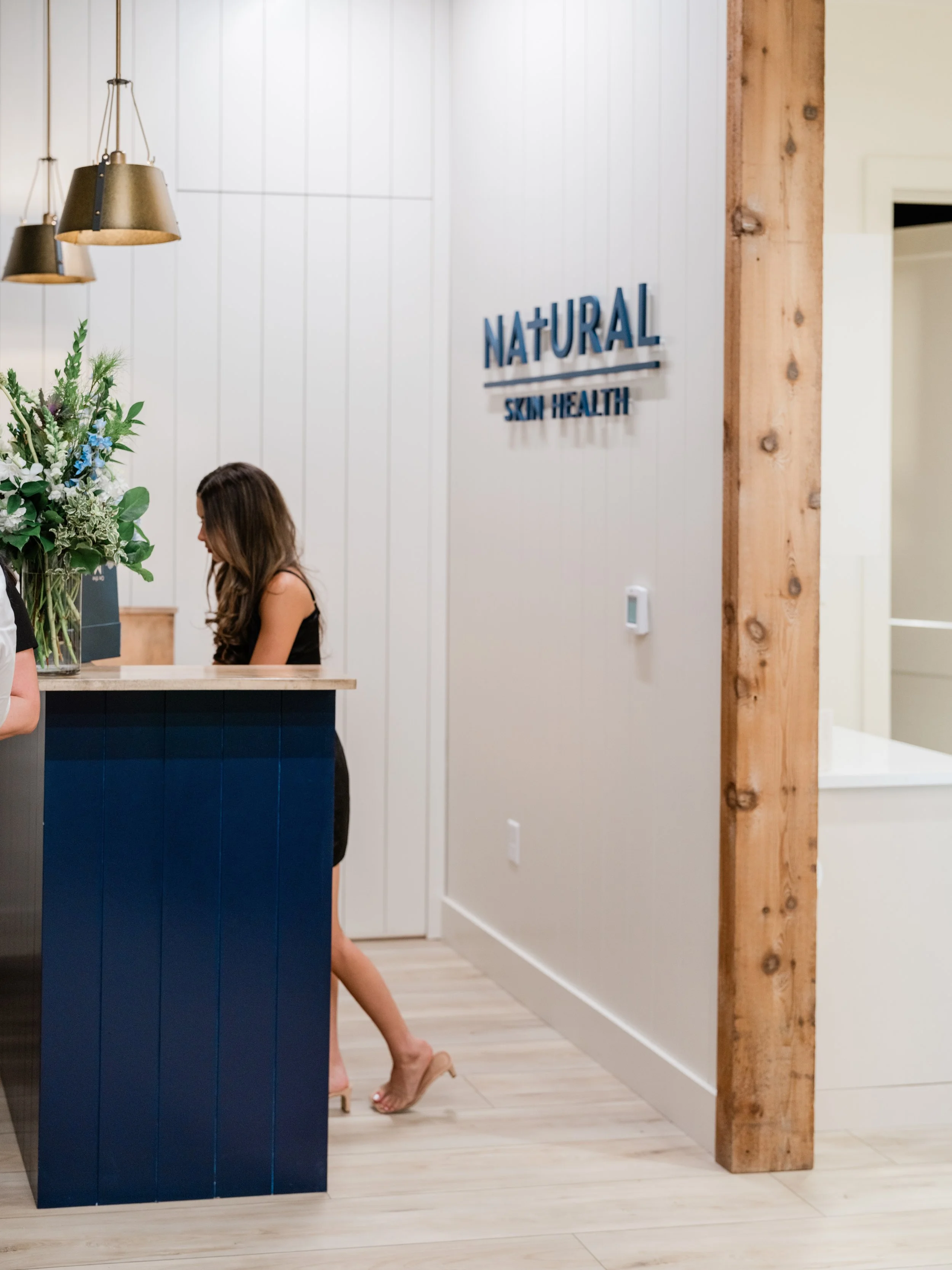 Natural Skin Health check-in desk. There are two gold pendants that hang over a navy blue reception desk. The Natural Skin Health logo is behind the desk.