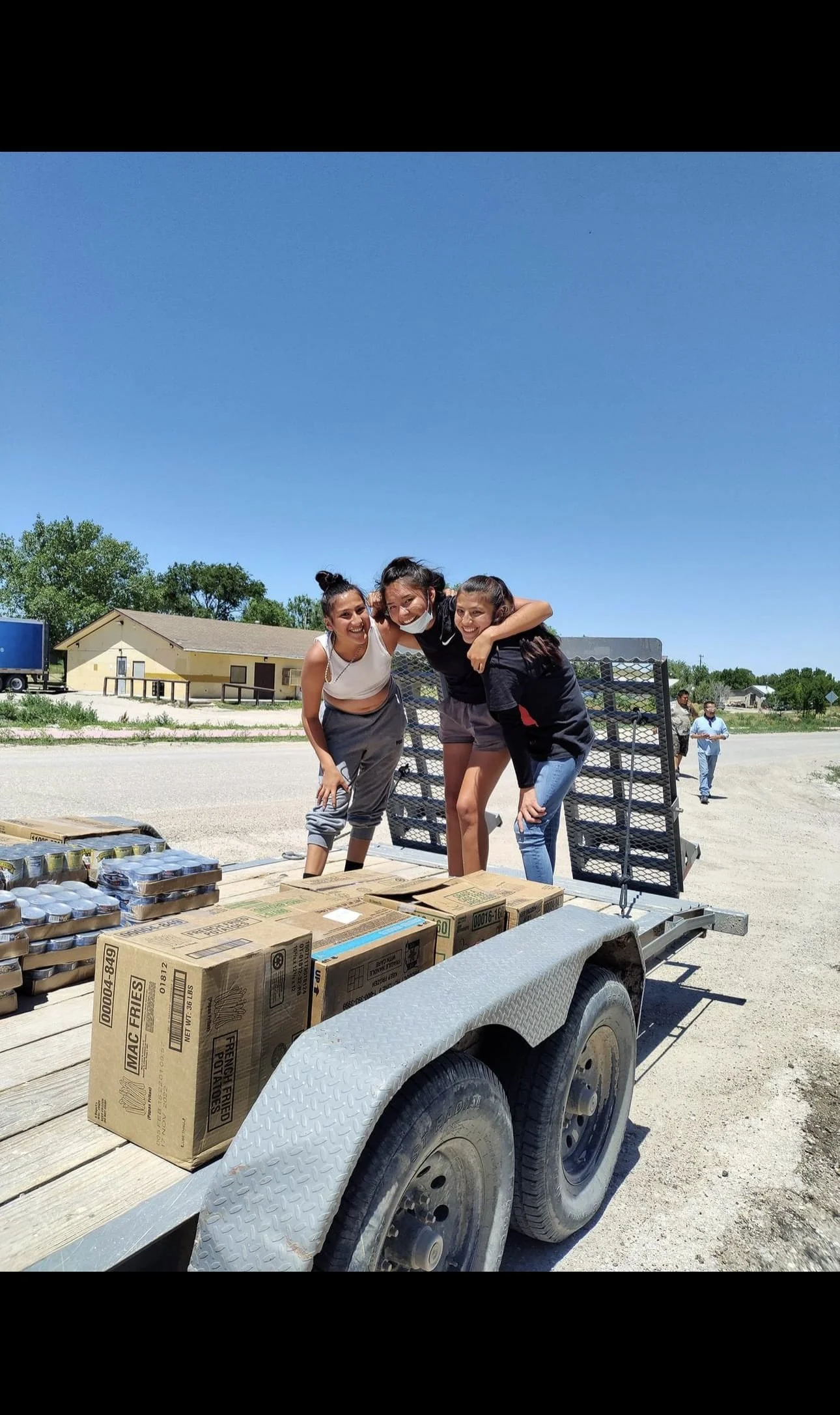 Three young women smiling and hugging on a trailer loaded with boxes and canned food outdoors under a clear blue sky.