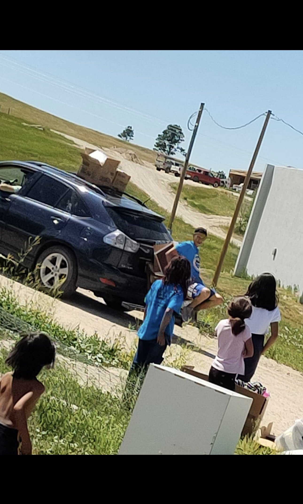 Children gathering around a black car with a trunk full of boxes on a suburban street on a sunny day.