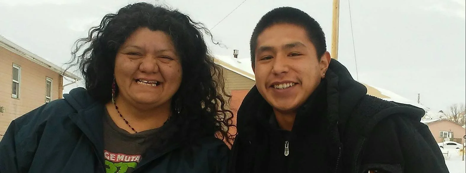 Two smiling people outdoors with cloudy skies, houses and a street in the background, wearing jackets.