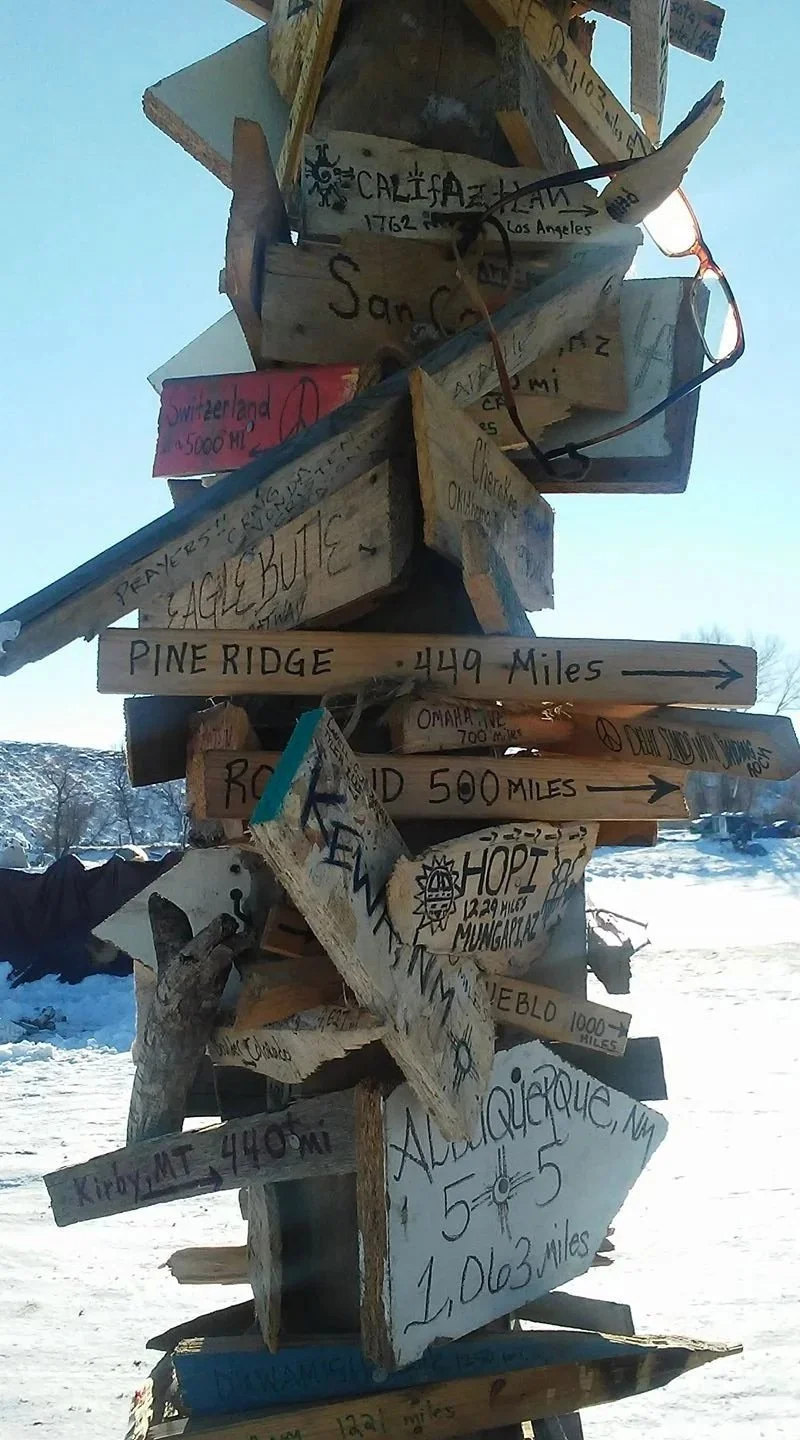 Signpost with multiple directional signs pointing to various locations, including Pine Ridge, Oklahoma, and Minnesota, with distances noted. The signs are handmade and stacked on a wooden pole in a snowy outdoor setting.