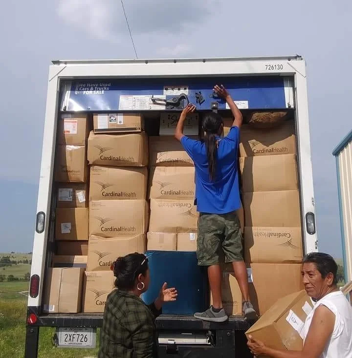 Children and adults unloading donated boxes from a truck labeled 'Cardinal Health,' with one child reaching up to adjust the truck's upper storage area.
