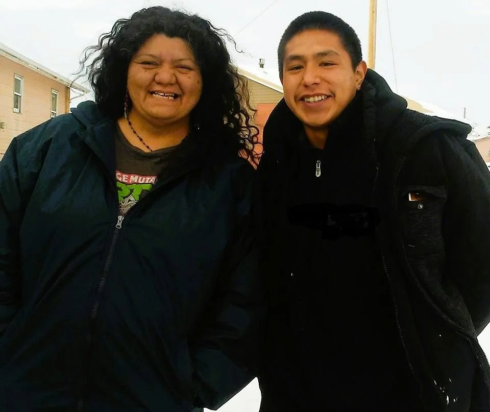 Two people smiling outdoors on a cloudy day, wearing dark jackets, with a row of houses in the background.