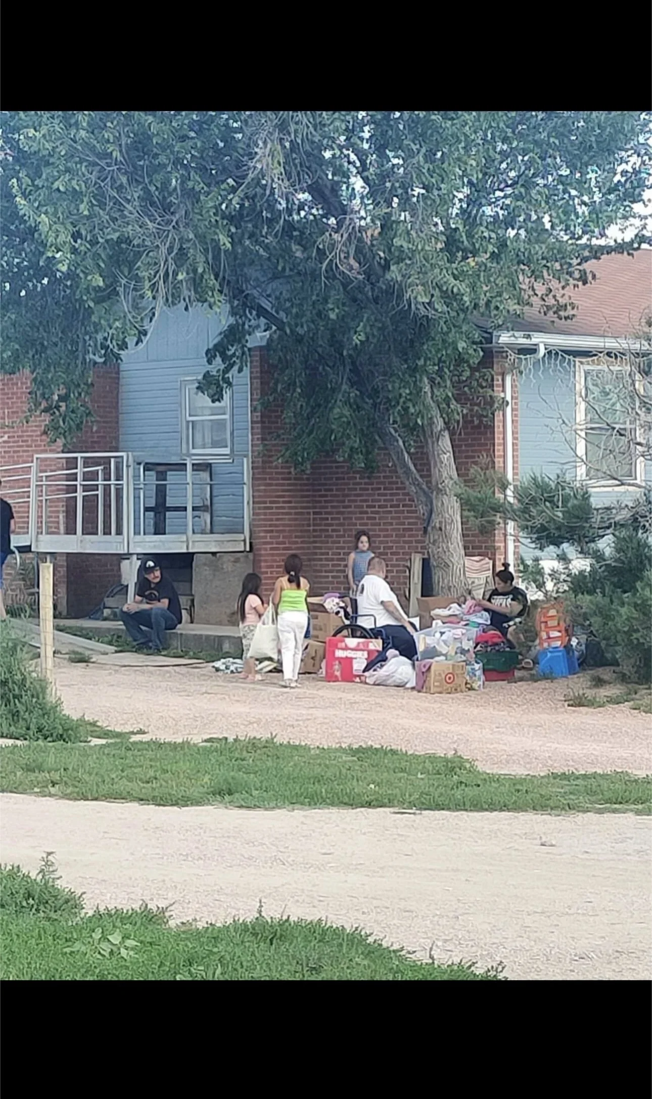 People gather outside a brick and siding house with emergency or community supplies, some sitting and others standing under a large tree.