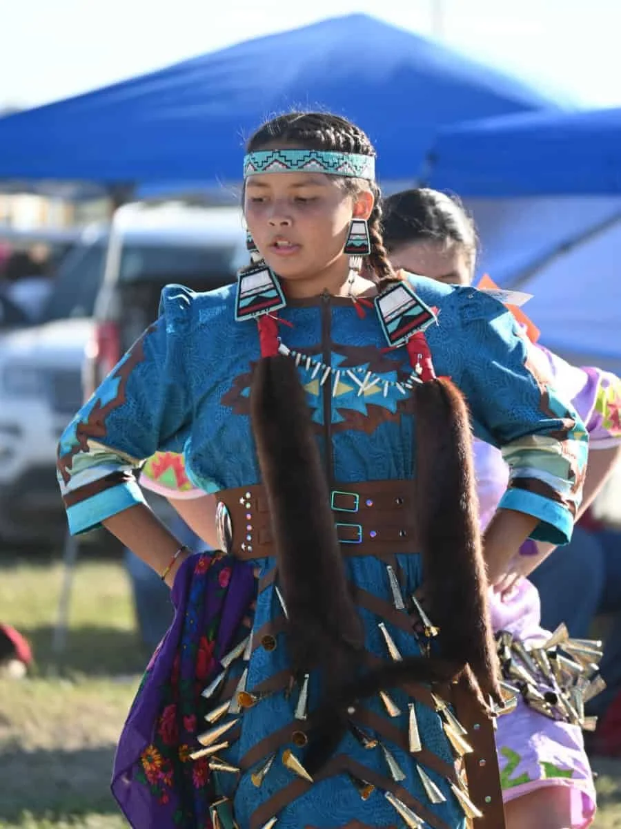Young girl dressed in traditional Native American attire with jewelry, standing outdoors with a blue tent and cars in the background.