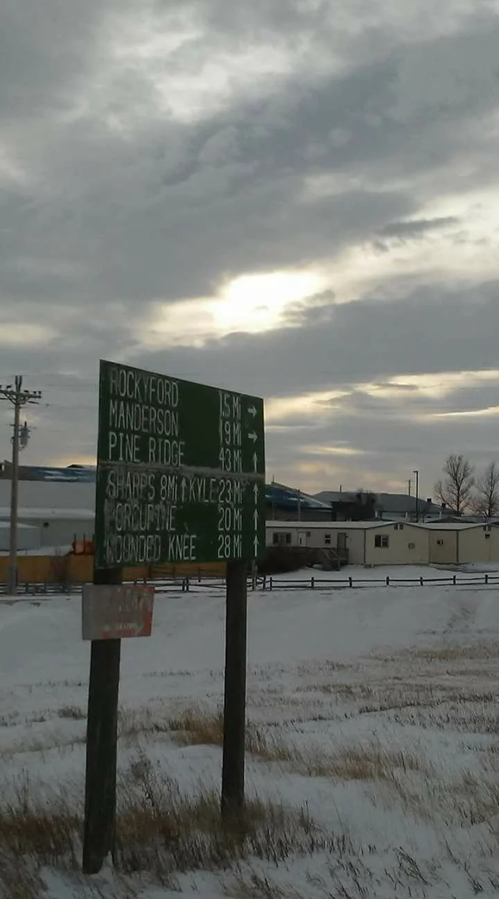 A road sign showing distances to Rocky Ford, Manderson, Pine Ridge, Sharp's, Crowline, and Mounted Knee in a snowy landscape with cloudy sky and some structures in the background.