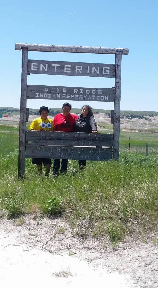 Three people standing behind a wooden sign at Pine Ridge Indian Reservation, with green grass and hills in the background.