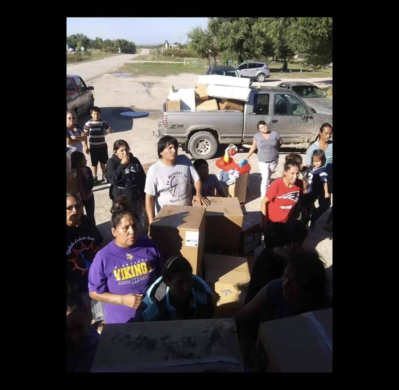 Group of people gathered outdoors with boxes and a pickup truck in the background, possibly distributing supplies.