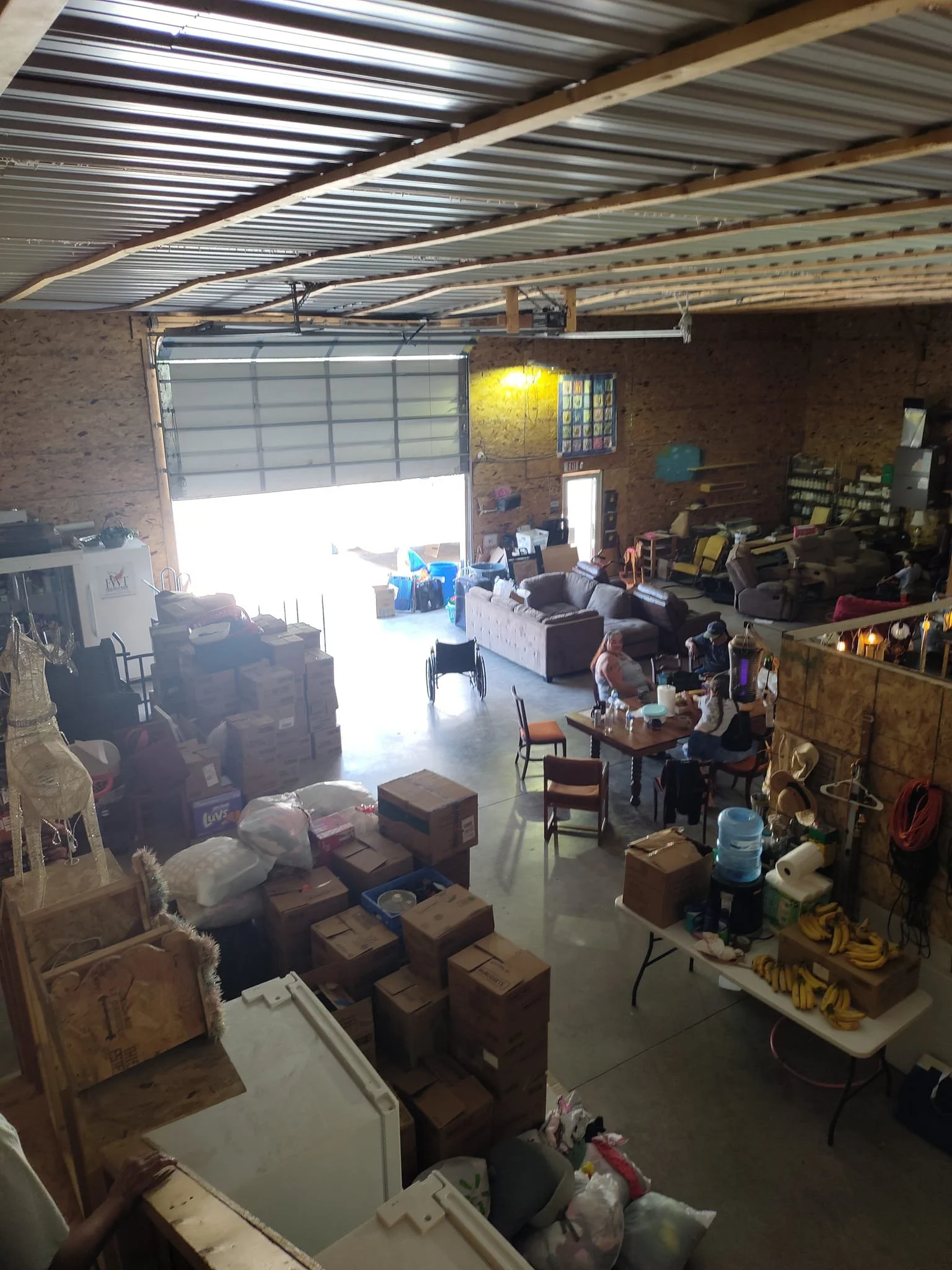 Interior of a warehouse or large storage space with boxes, furniture, and people sitting at tables.