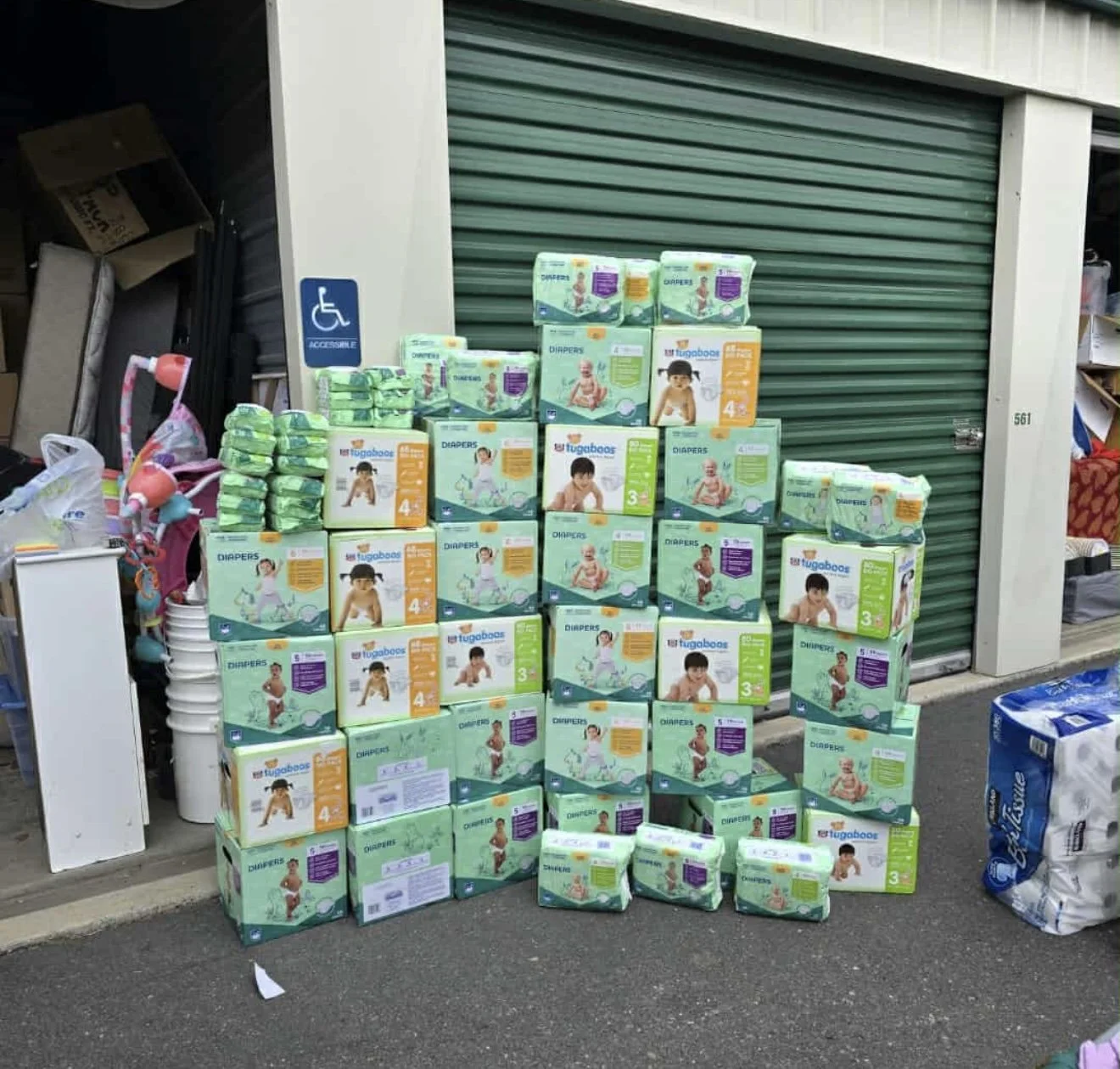 Stacked boxes of baby diapers and wipes outside a store with a green roll-up door, alongside a display of colorful children's toys.