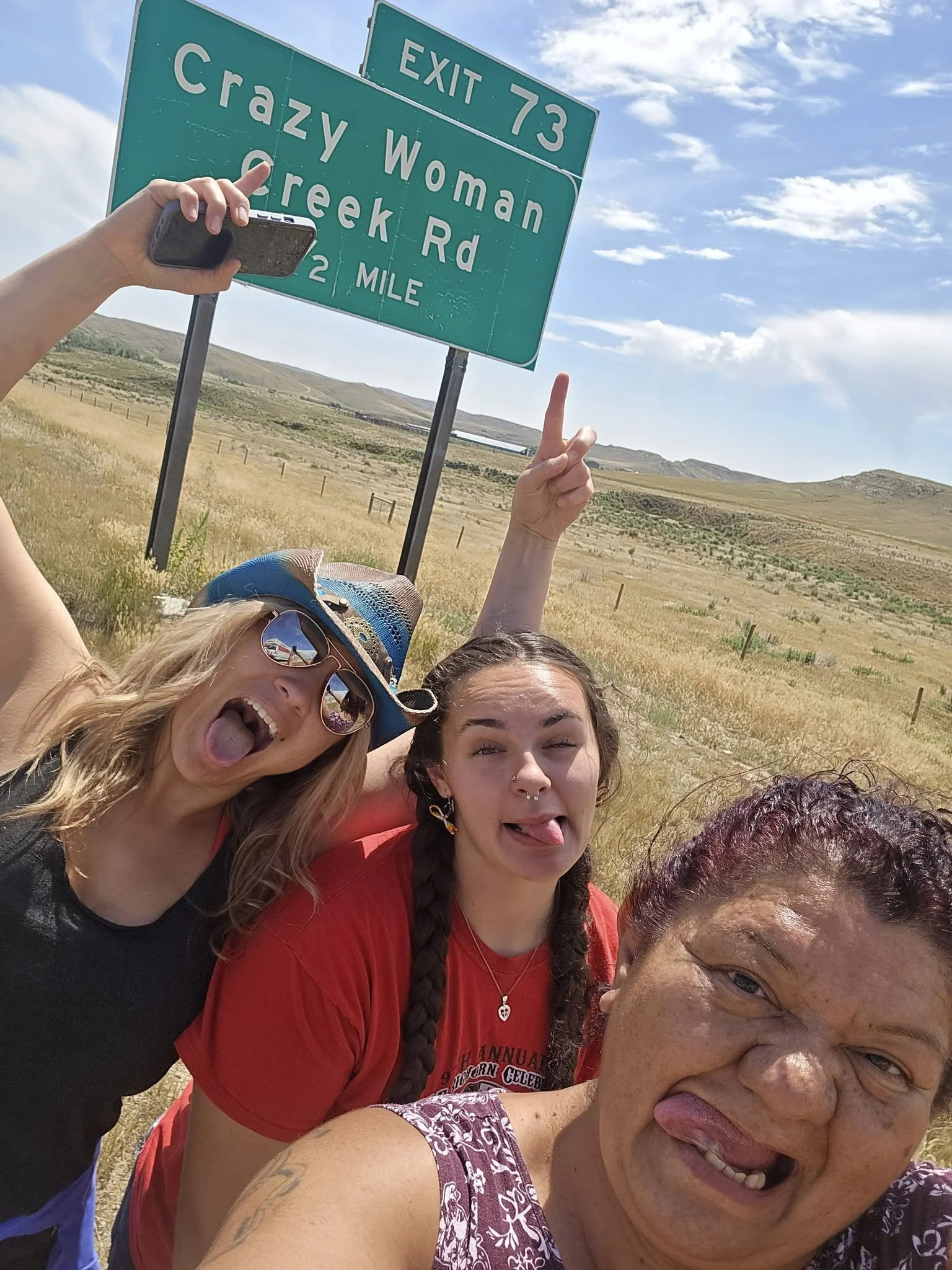 Three women taking a selfie in front of a green highway sign that reads 'Exit 73 Crazy Woman Creek Rd, 2 Mile'. They are sticking out their tongues and making faces, with a rural landscape in the background.