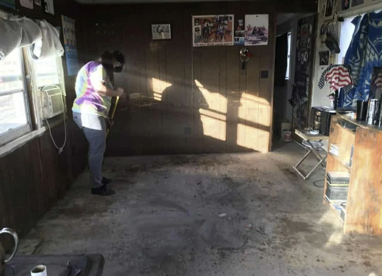 Person cleaning the floor with a broom in a rustic indoor space with wood-paneled walls, an air conditioning unit in the window, and various decorations including pictures on the wall.