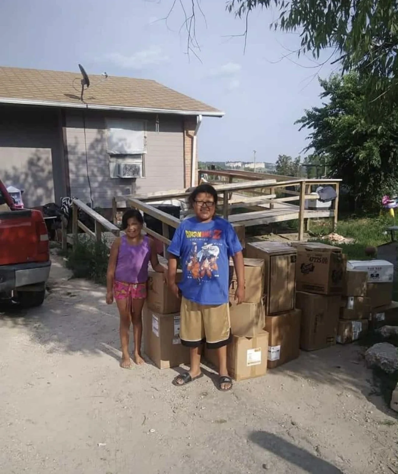 Two children standing in front of a pile of cardboard boxes on a dirt road, outside a house with a ramp, trees, and a parked red vehicle.