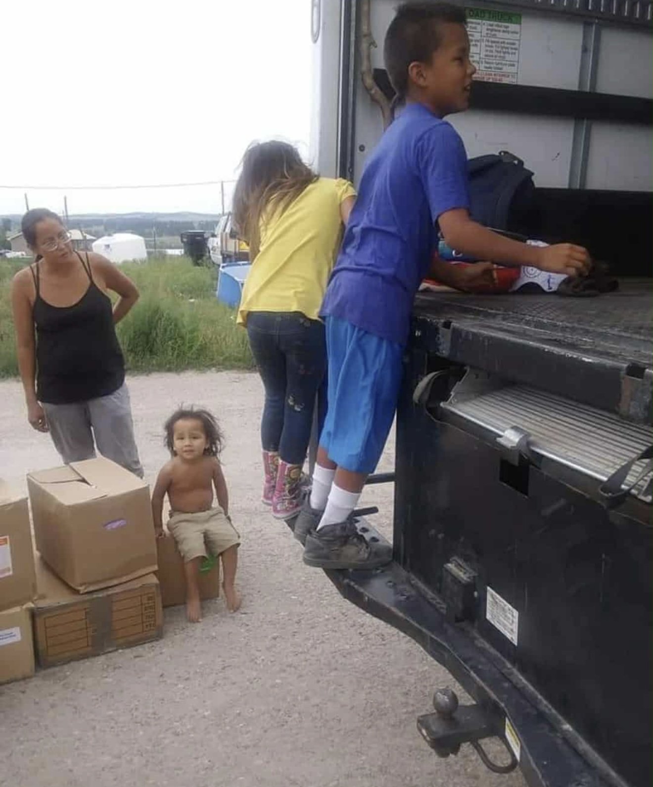 Two children jump onto the back of a truck, while three women observe. One young girl sits on boxes nearby, and a woman stands with her hands behind her back. The setting appears to be outdoors on a gravel lot with some grassy fields in the backgroun