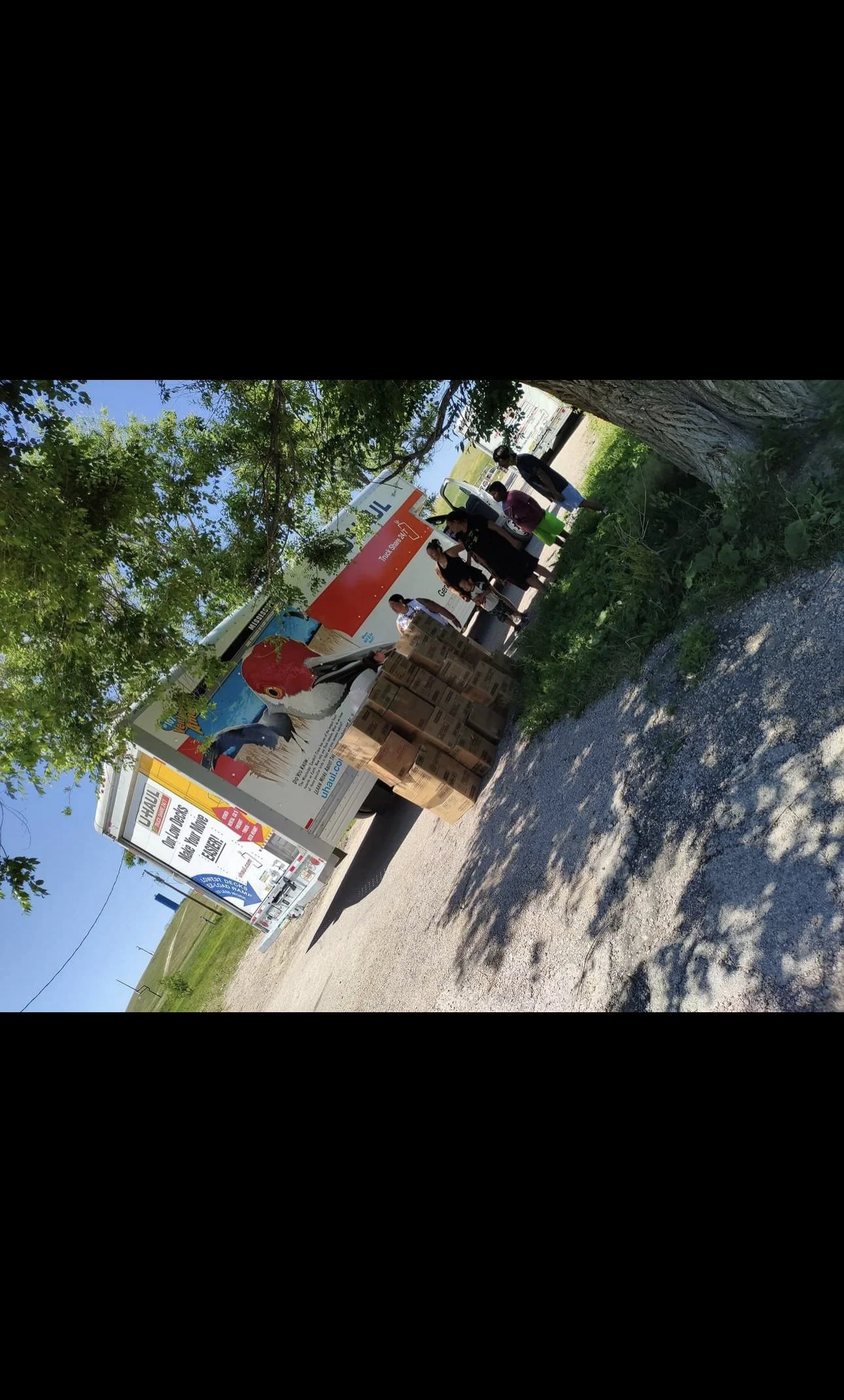 People standing around a food truck with a large image of a chicken on its advertisement, and boxes stacked on the ground nearby, under trees on a sunny day.