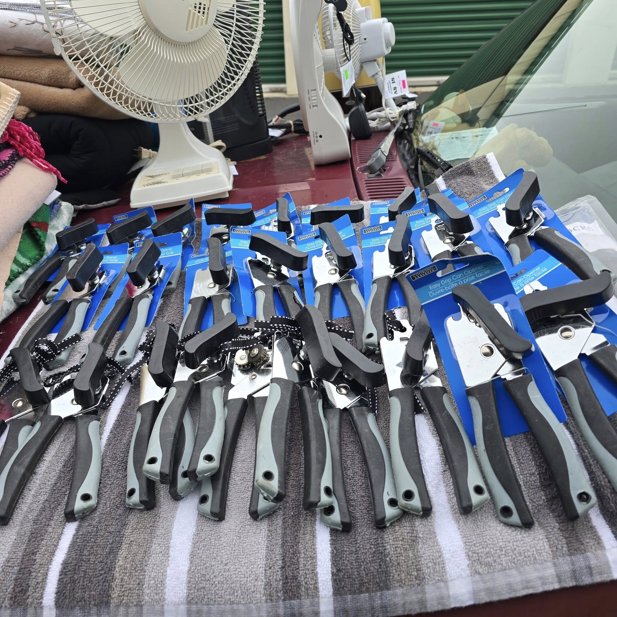 A display of multiple black and gray can openers on a striped towel at a market stall, with a fan, electronics, and other items in the background.