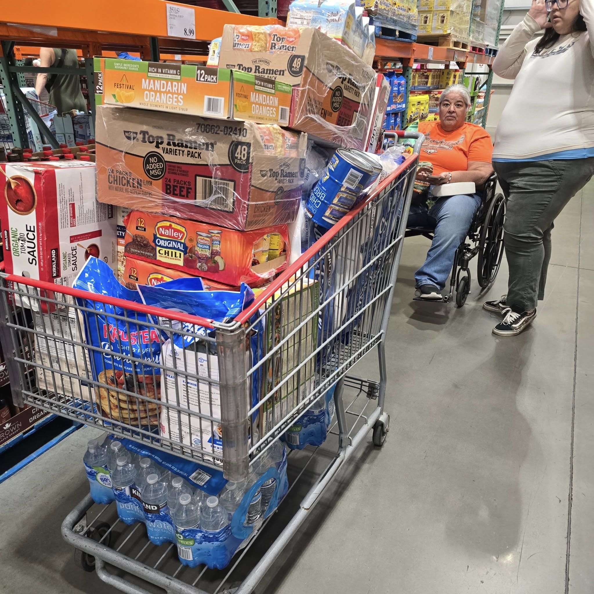 A shopping cart filled with various grocery items, including bottled water, ramen packs, chili seasoning, and boxes of oranges, inside a store aisle. In the background, a woman in a wheelchair and another woman are shopping.