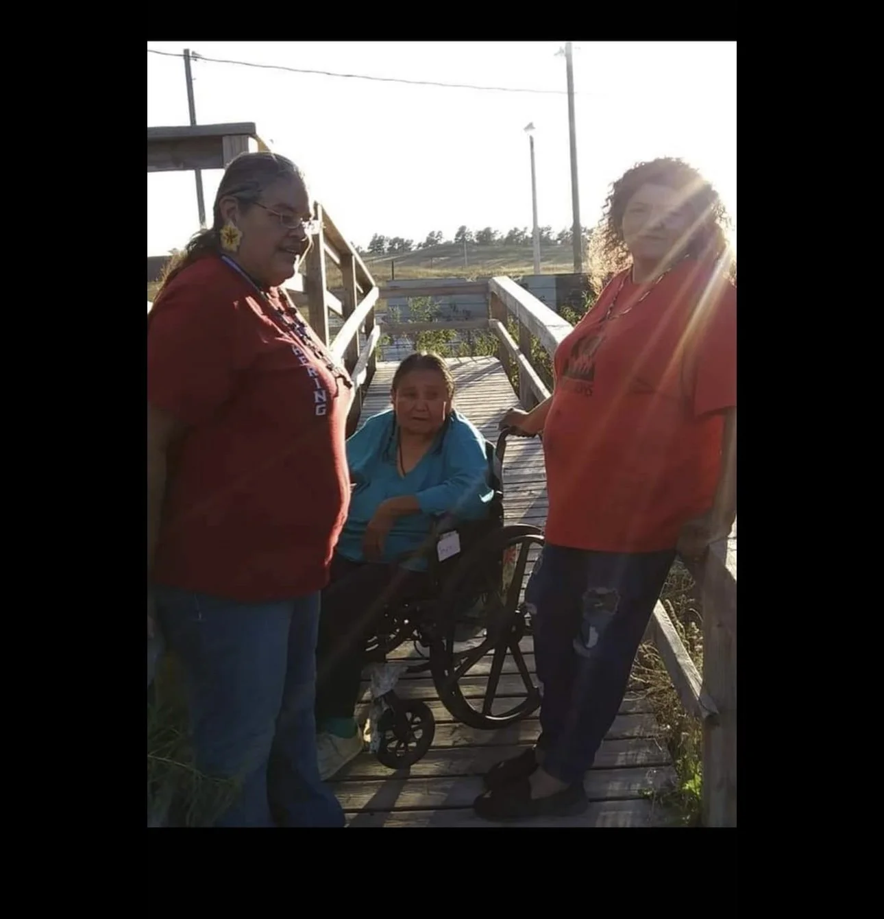 Three women, one in a wheelchair, standing on a wooden bridge outdoors in sunlight.