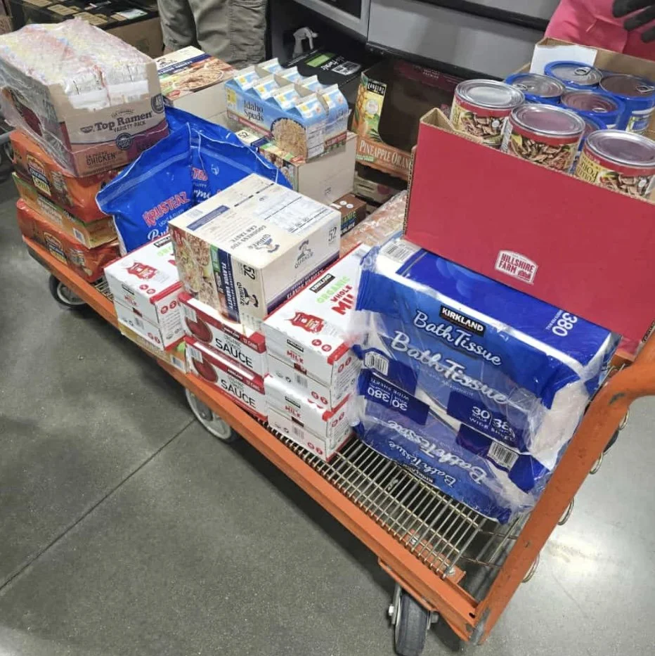 A shopping cart filled with various grocery items, including canned goods, boxes of tissues, ramen noodles, and other packaged food products.