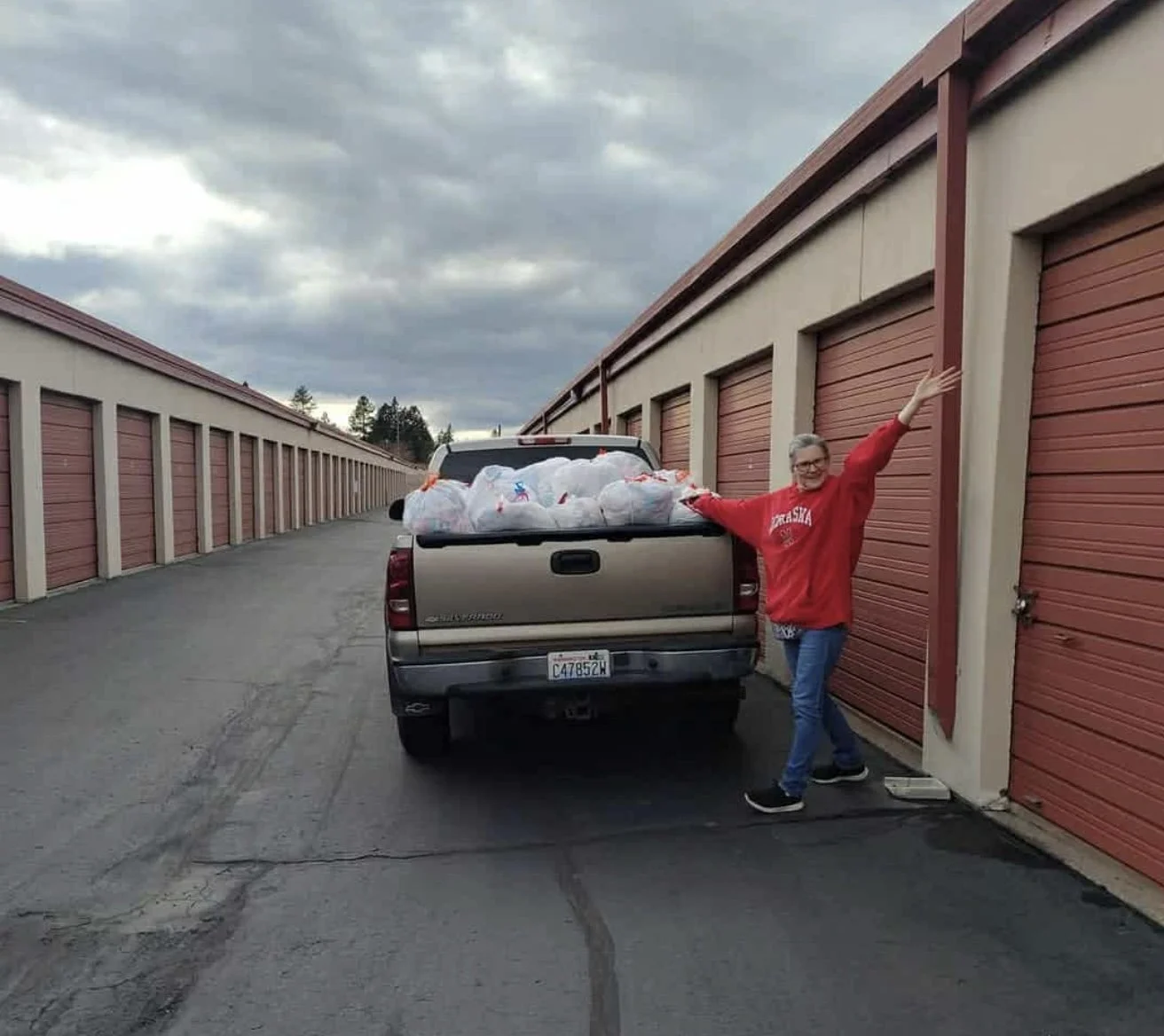 A man standing beside a pickup truck filled with garbage bags in a storage unit parking area, with his arm raised and a smile on his face.