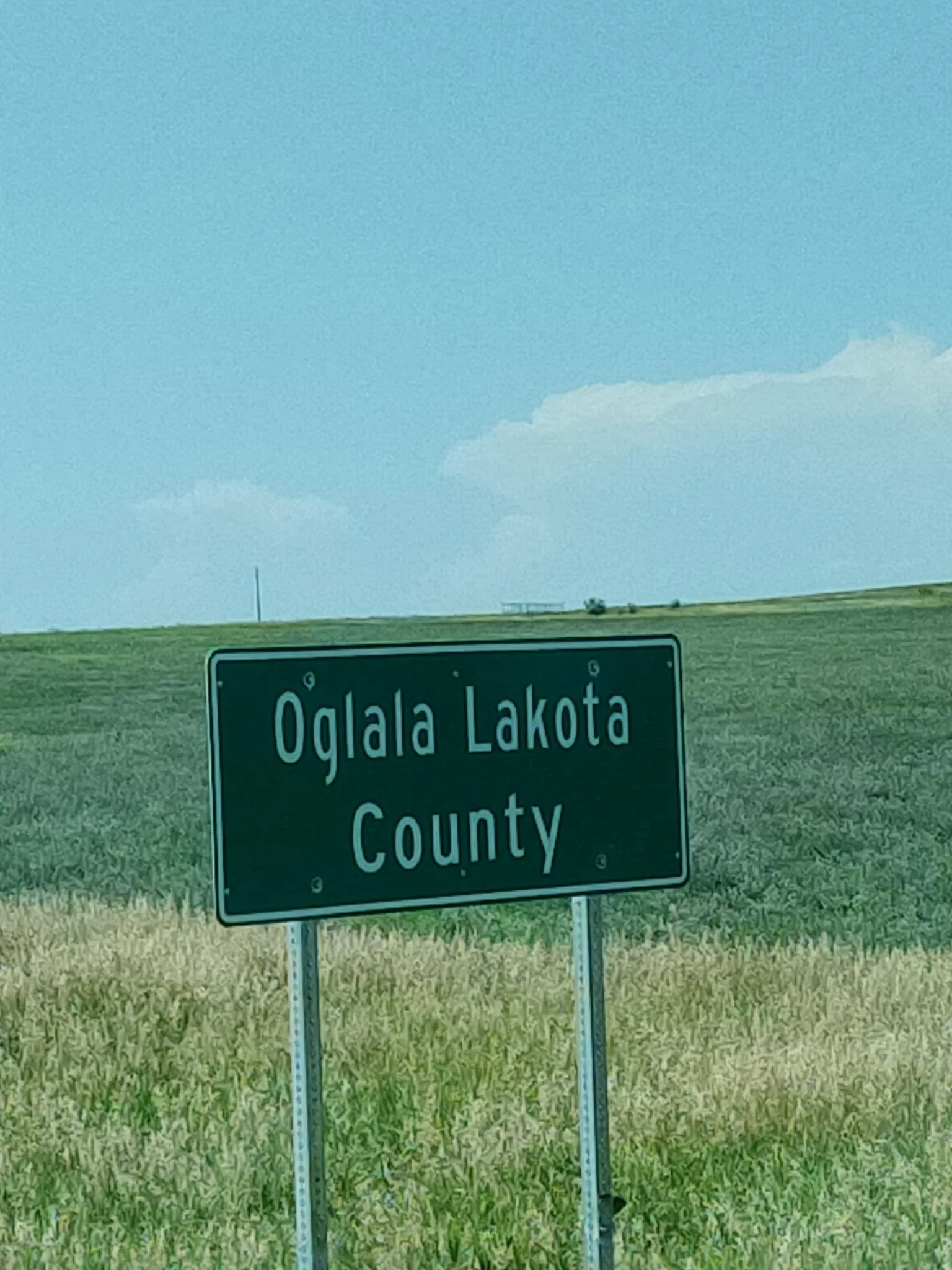Green road sign indicating Oglala Lakota County with a grassy field and blue sky in the background.