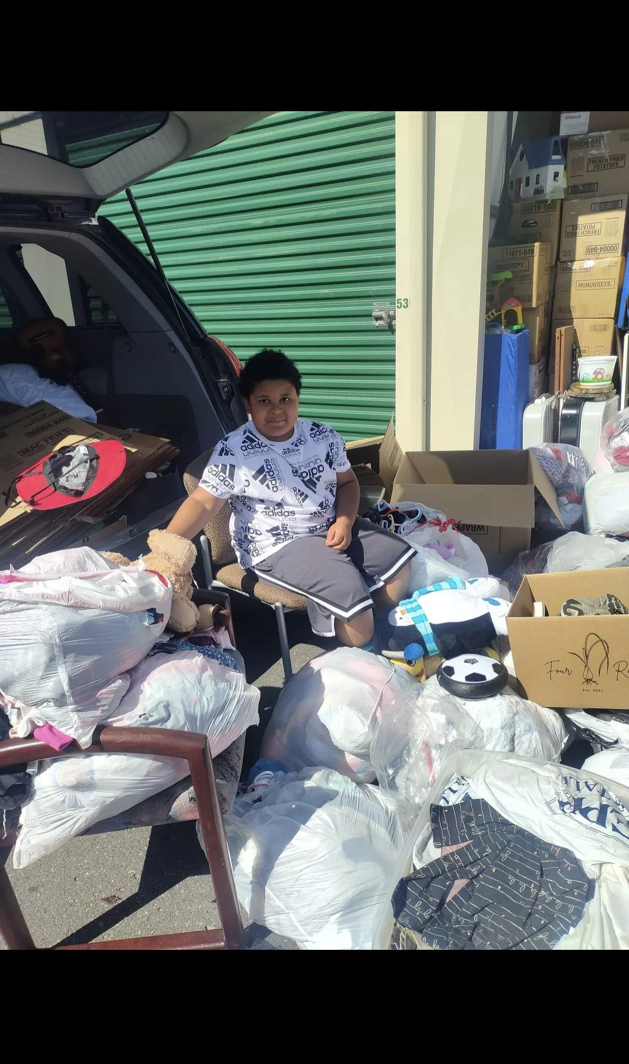 A young boy sitting on a chair surrounded by bags and boxes of clothes and toys at a yard sale or flea market, with a partially opened van and storage area in the background.