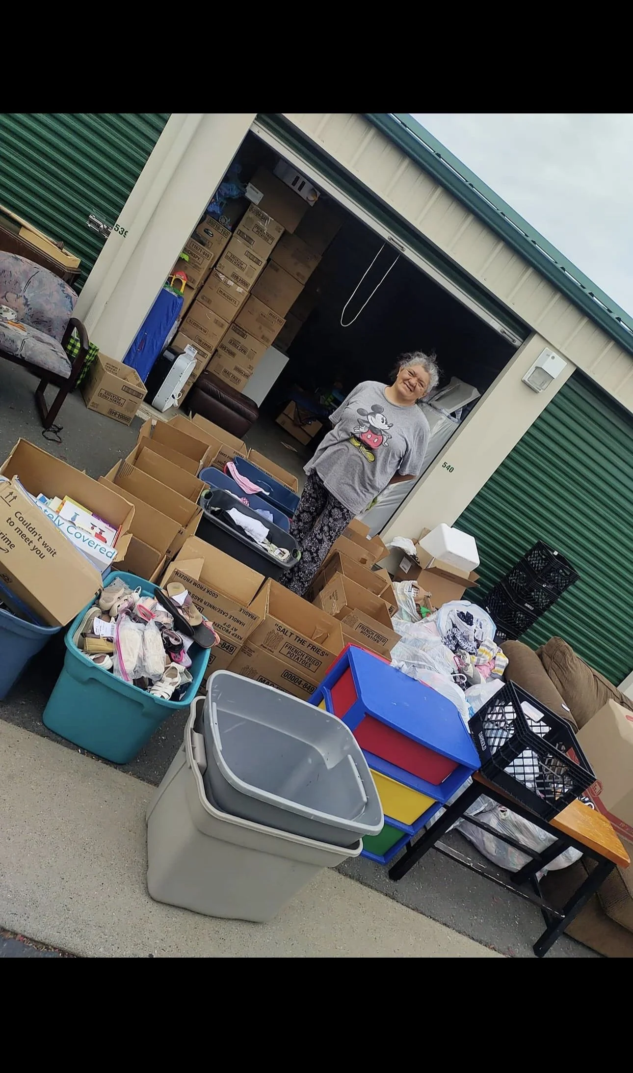 A woman standing behind tables and boxes filled with various items like shoes, books, and miscellaneous household goods outside a storage unit or shop.