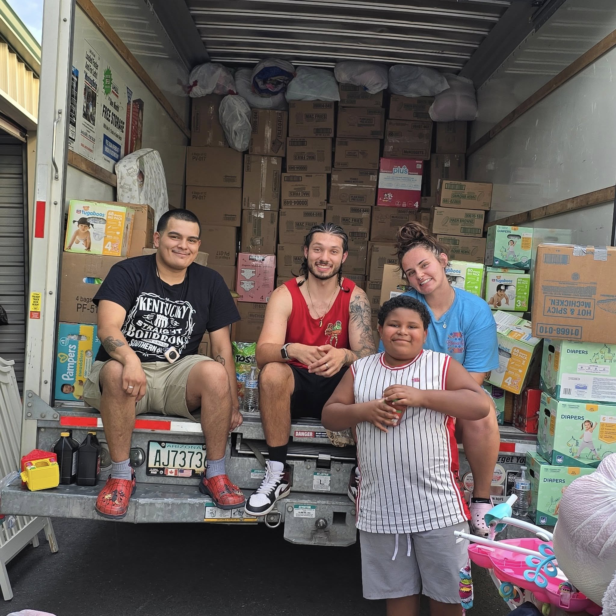 People sitting inside and around a truck loaded with boxes of donated supplies, including diapers and baby products. The group consists of two adults and two children, smiling for the camera.