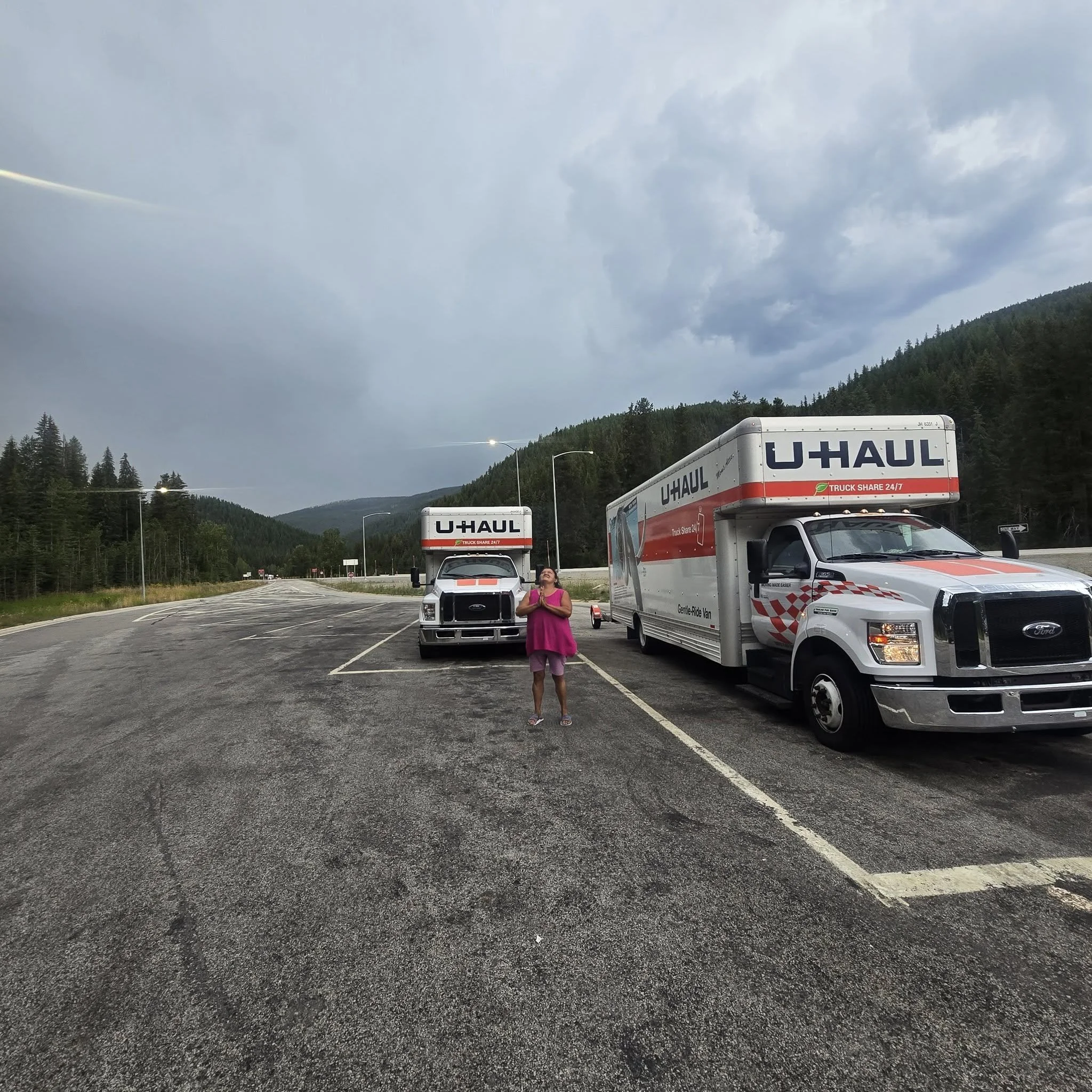 A woman in a pink dress standing in an empty parking lot with two U-Haul trucks parked nearby, surrounded by green mountains and cloudy sky.