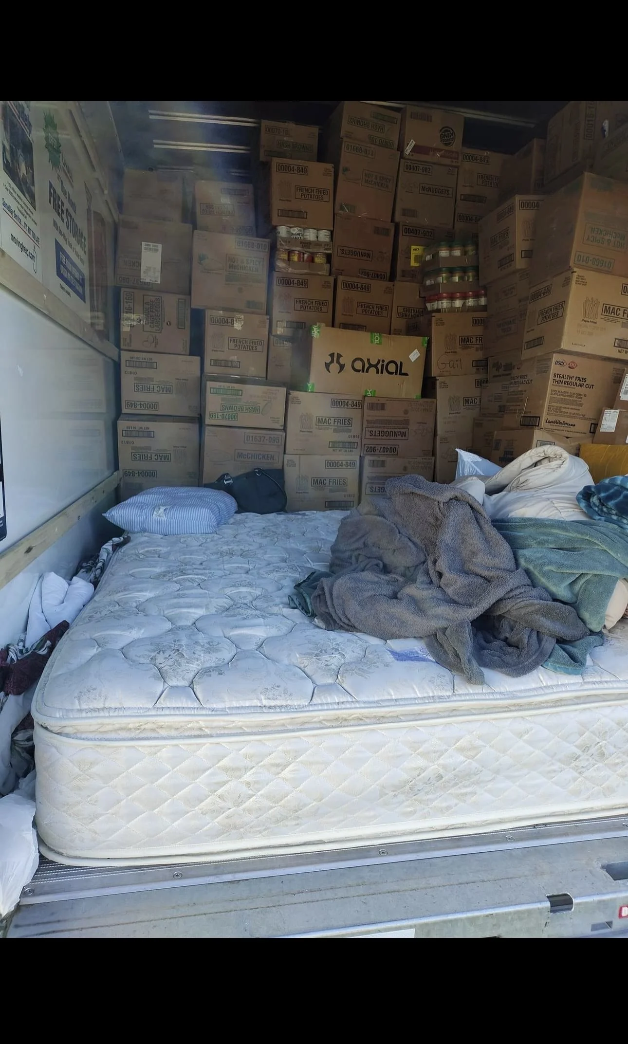 An inside view of a storage space or moving truck with a mattress, some blankets, a pillow, and a black bag on top, surrounded by stacked boxes filled with food and supplies.