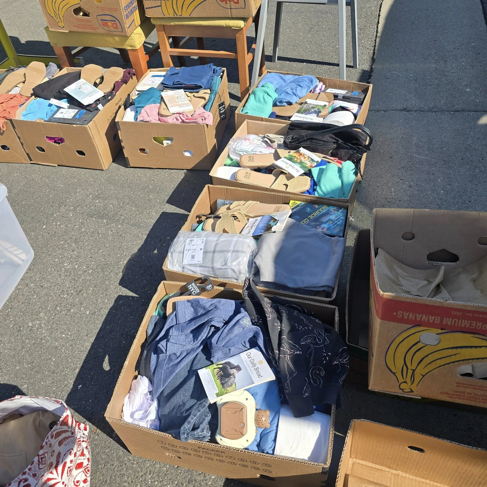 Boxes filled with various clothing, shoes, and household items at an outdoor flea market or swap meet.