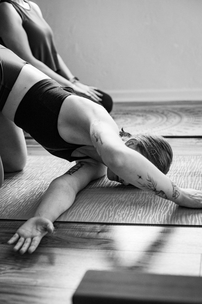 A group of women practicing yoga in a bright, spacious room with wooden floors and large windows, using yoga mats and blocks.