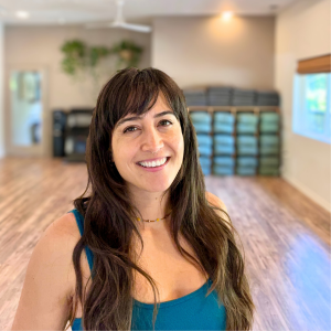 A young woman with long, wavy hair smiling at the camera in a room with a blurred background and green plants.