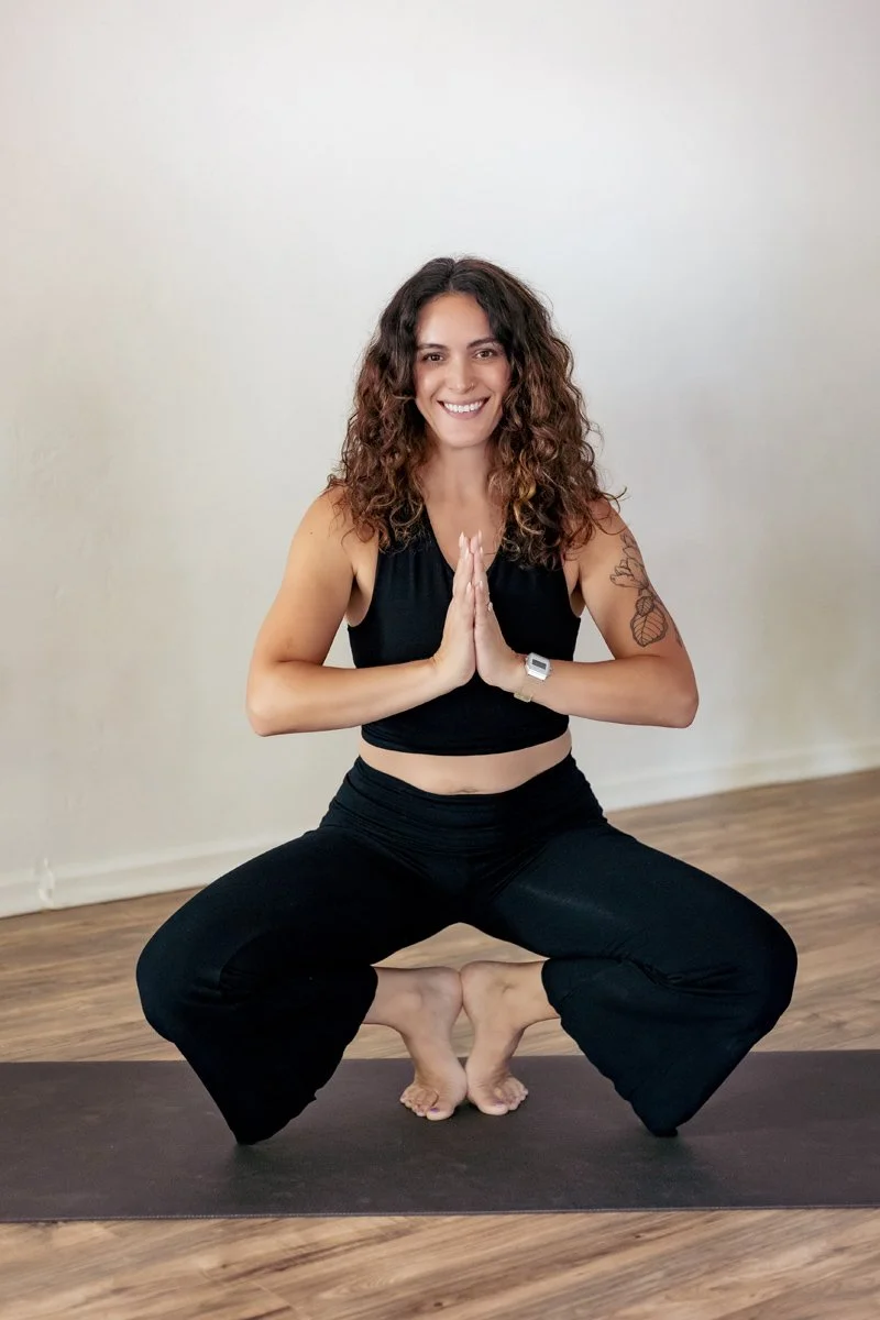 A woman with curly brown hair smiling, wearing a black sleeveless crop top, sitting indoors with a blurred background of wooden flooring and furniture.