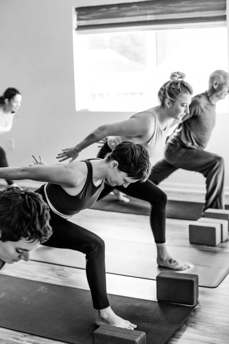 A woman practicing yoga indoors, standing on a yoga mat with arms extended and legs apart, in a well-lit room with wooden floors and potted plants.
