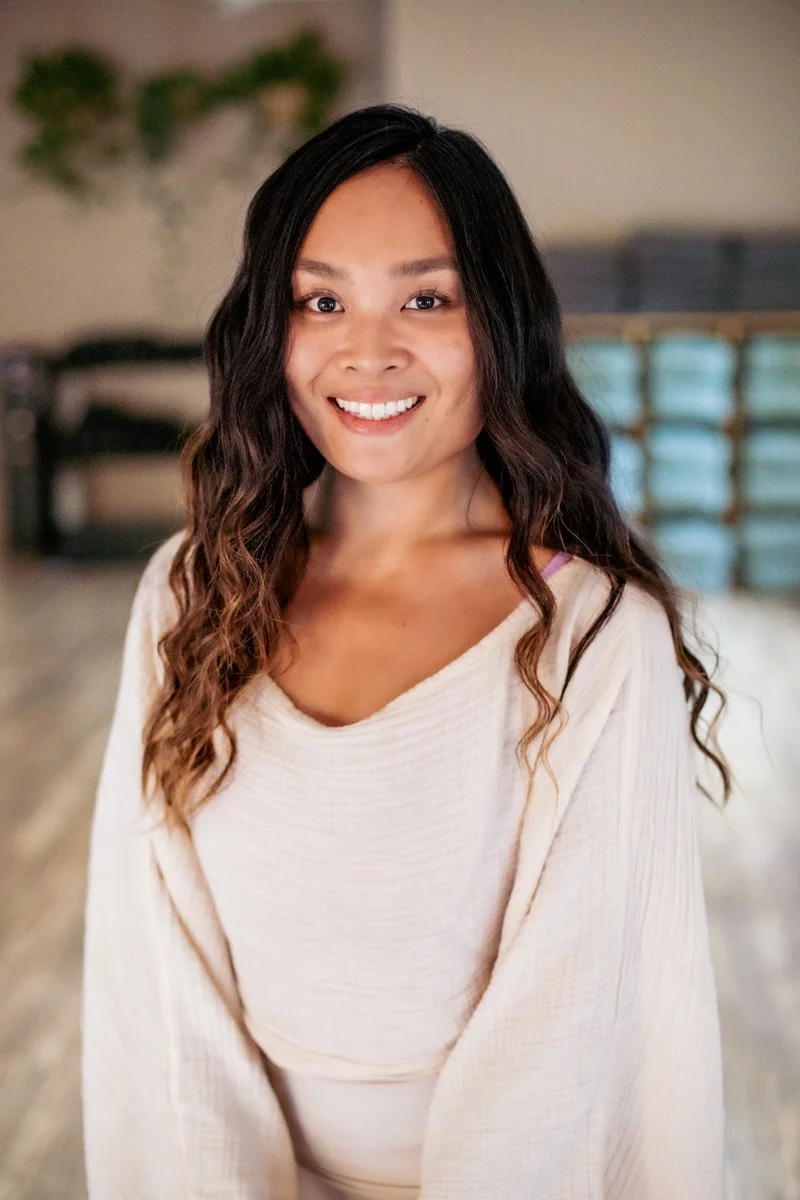A young woman with long, wavy hair smiling at the camera in a room with a blurred background and green plants.
