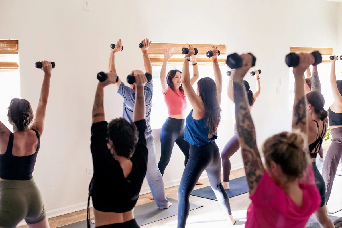 A pair of black dumbbells labeled 8 pounds on a wooden gym floor, with multiple similar dumbbells stacked against a wall in the background.
