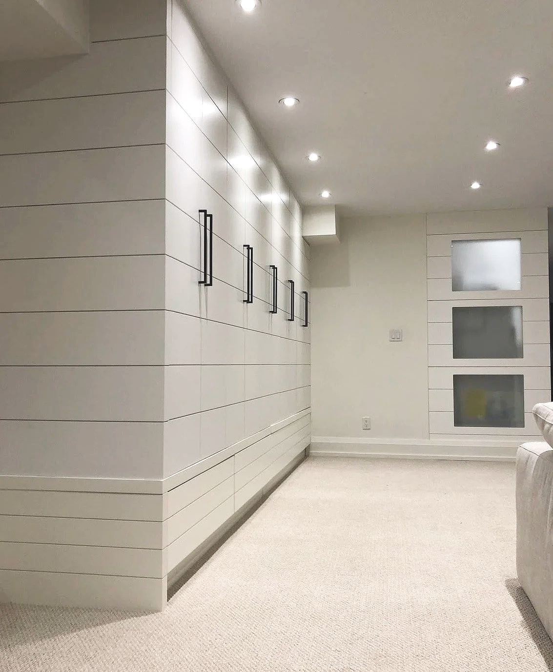A modern storage room with white walls, black door handles, a row of small ceiling  pot lights, a small window with frosted glass on the right, and light-coloured carpeted flooring.